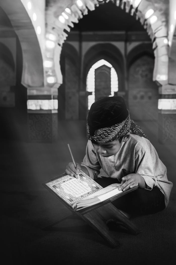 A young child deeply engaged in reading the Quran inside a mosque, captured in black and white.